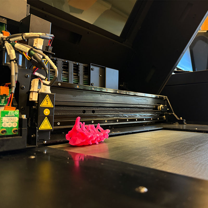 A 3D printer in operation producing a bright pink, organic-shaped model on the print bed, with visible wiring and safety warning labels on the machine.