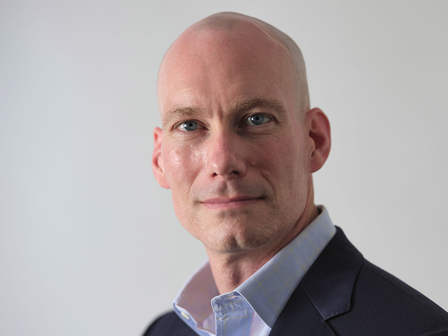 Studio headshot of a bald white man wearing a dark blazer and light blue collared shirt, photographed from the chest up against a light gray background, facing slightly to the side with direct eye contact.