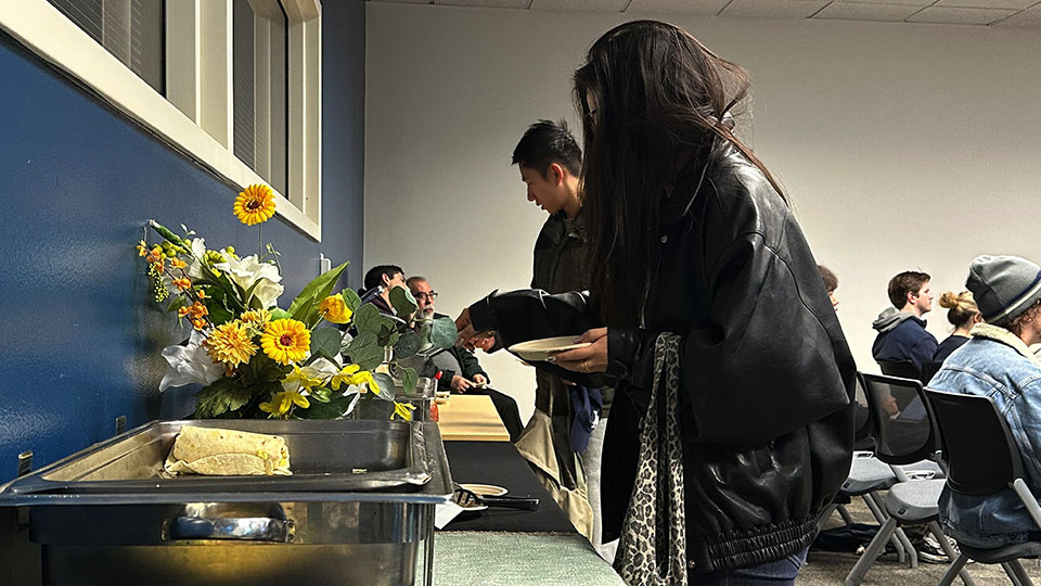 Person serving themselves food from a buffet table adorned with a floral arrangement while others sit and talk in the background of a classroom-style setting.