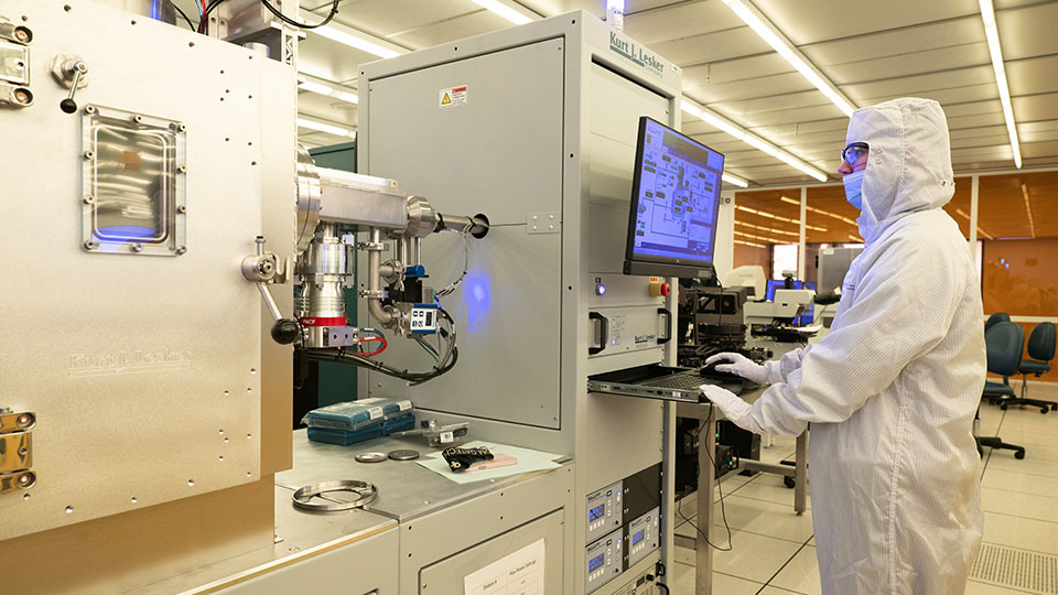 Person in a cleanroom suit operating a high‑precision laboratory instrument with a computer interface.