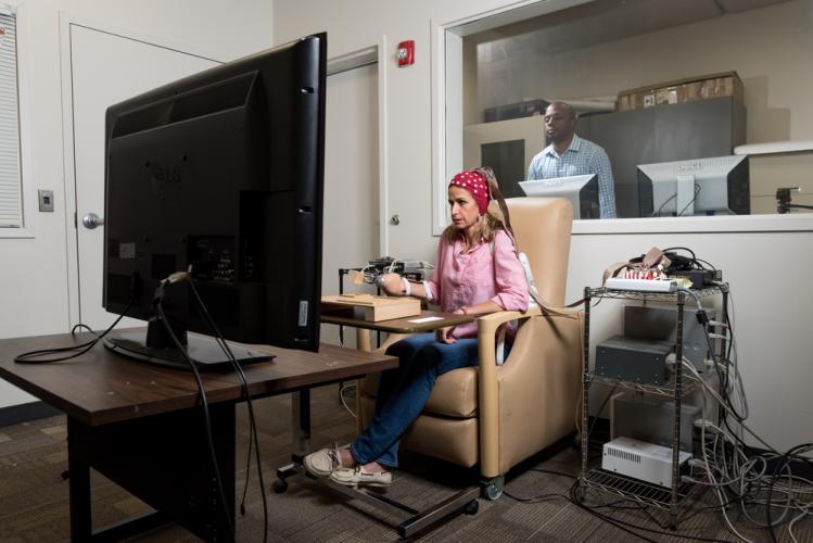 A person seated in a beige chair using a computer setup with multiple cables and devices, facing a large monitor in a testing or research room, with another individual visible through a window in an adjacent control room.